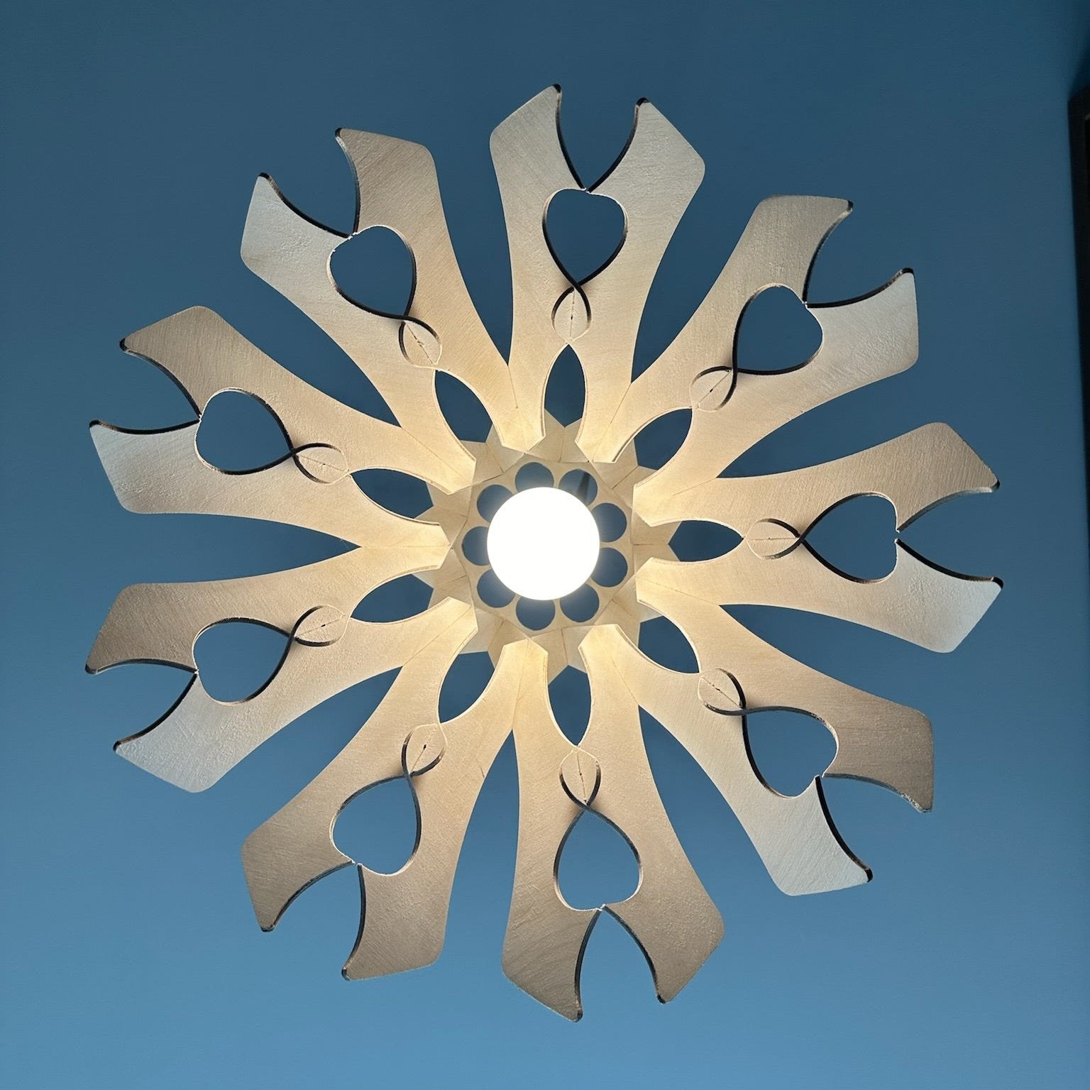 Bottom view of a natural birch wood pendant lampshade with heart-shaped cutouts. The light shines through the design, casting a soft, warm glow and creating a pattern on the ceiling.