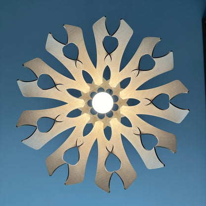 Bottom view of a natural birch wood pendant lampshade with heart-shaped cutouts. The light shines through the design, casting a soft, warm glow and creating a pattern on the ceiling.