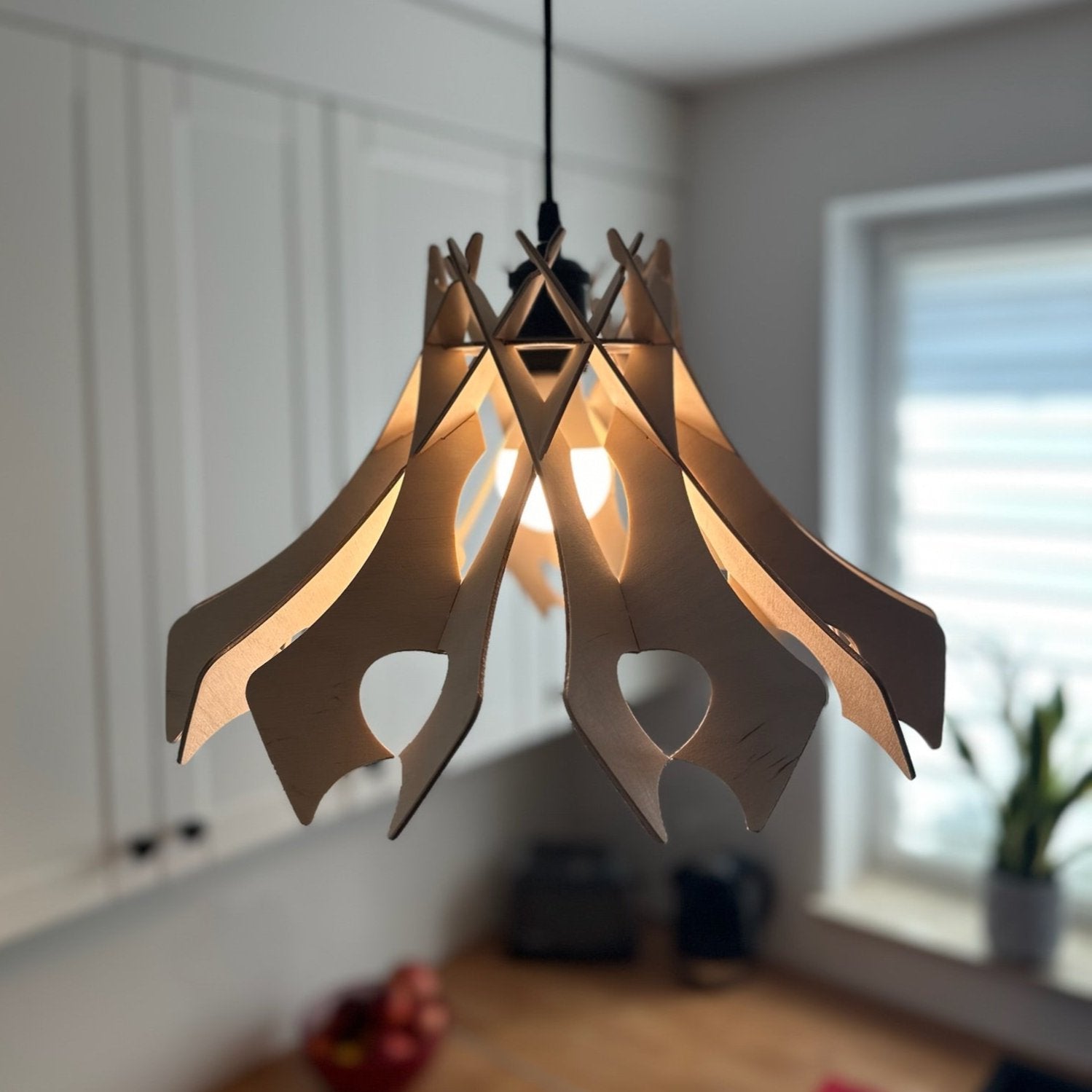 Natural birch wood pendant lampshade with smooth, curved cutouts, hanging in a contemporary kitchen. The lampshade emits a soft, warm light, complementing the white and wooden decor.