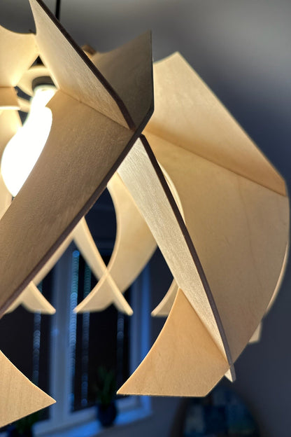 Close-up of the edge of a large wooden pendant lamp, showcasing the smooth, curved design and natural birch wood texture. The warm light from the bulb highlights the precise craftsmanship and modern aesthetic.