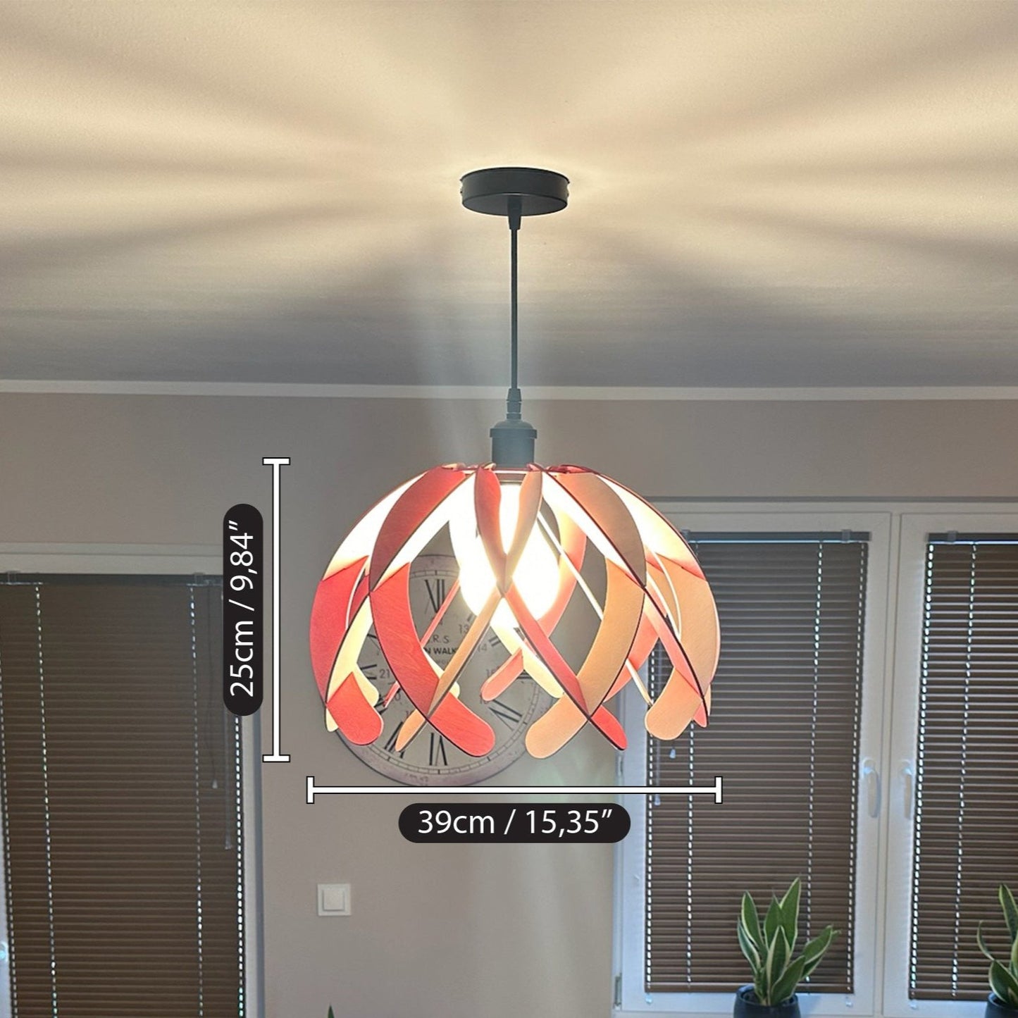 Wooden pendant lamp in white and pink, hanging from the ceiling with a black cord. The dimensions of the lampshade are shown: 25cm (9.84 inches) in height and 39cm (15.35 inches) in diameter. The light pattern casts beautiful shadows on the ceiling.