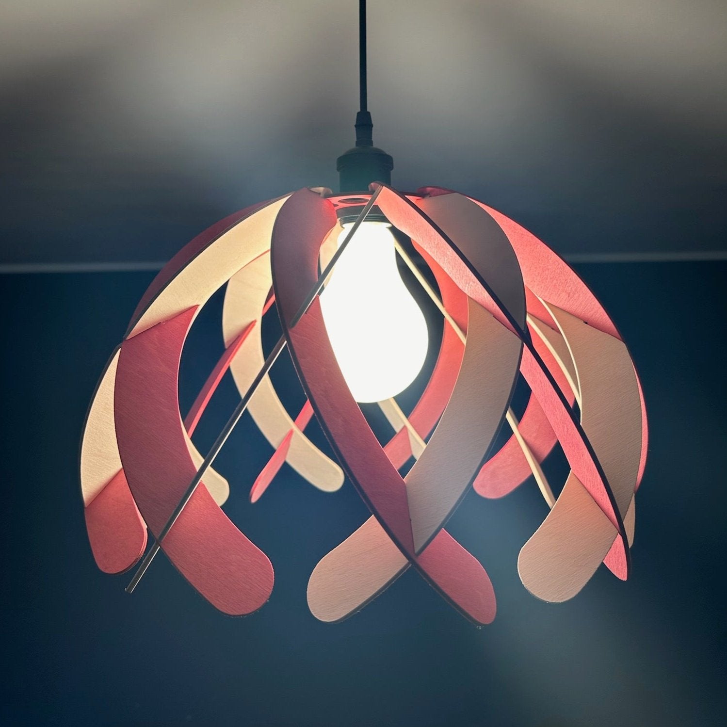 Close-up of the pink and white wooden pendant lampshade with the light turned on, revealing the smooth finish and detailed wooden design. The black socket and cord are visible, enhancing the modern aesthetic.